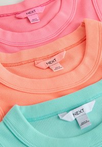 Trois t-shirts côtelés empilés : le dessus en rose clair, le milieu en corail, le dessous en vert menthe, tous avec des étiquettes indiquant la marque "NEXT" et l'origine "fabriqué au Bangladesh".