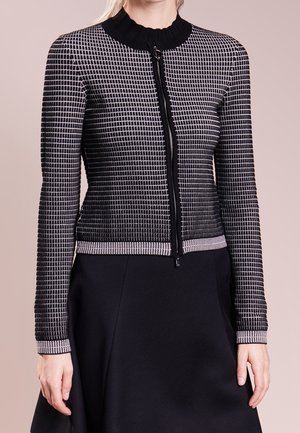 Woman wearing a black and white checkered zip-up cardigan with ribbed collar and cuffs, paired with a black skirt.