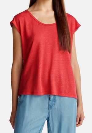 Kvinde iført en løs rød top med kappelignende ærmer parret med lyseblå højtaljede denimjeans mod en hvid baggrund.