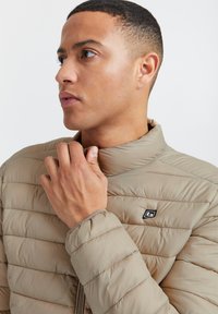 Chaqueta acolchada beige con cuello alzado, costuras horizontales y un parche de logo negro en el pecho. Material suave y ligero.