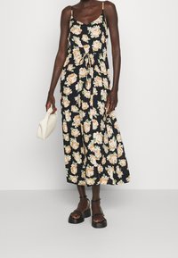 Květinové maxi šaty s černým pozadím, zdobené velkými broskvovými růžemi a zelenými listy, s nastavitelnými ramínky na spaghetti a detailem vázání v pase.