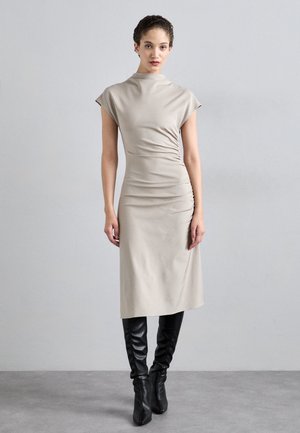 Beige muotopuvun midi-mittainen mekko, jossa on lyhyet hihat, kerätystä sivusaumasta yksityiskohtia, sileä pinta, yhdistettynä polven yli ulottuviin mustiin nahkabootseihin.