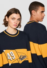 Granatowy sweter z żółtymi paskami i graficzną naszywką z numerem 49. Żebrowany dekolt i teksturowany materiał. Luźny krój.