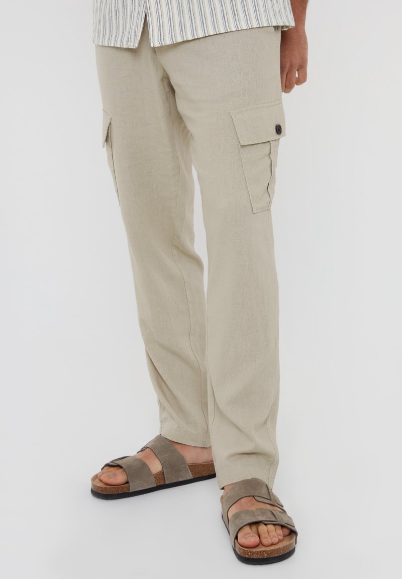 Pantalones cargo beige con bolsillos laterales abotonados, usados con sandalias de doble tira en tono topo sobre fondo blanco.