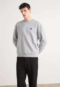 Sudadera de algodón gris con cuello redondo, puños y dobladillo acanalados, que presenta un pequeño parche de logo negro en el pecho. Combinada con pantalones negros.