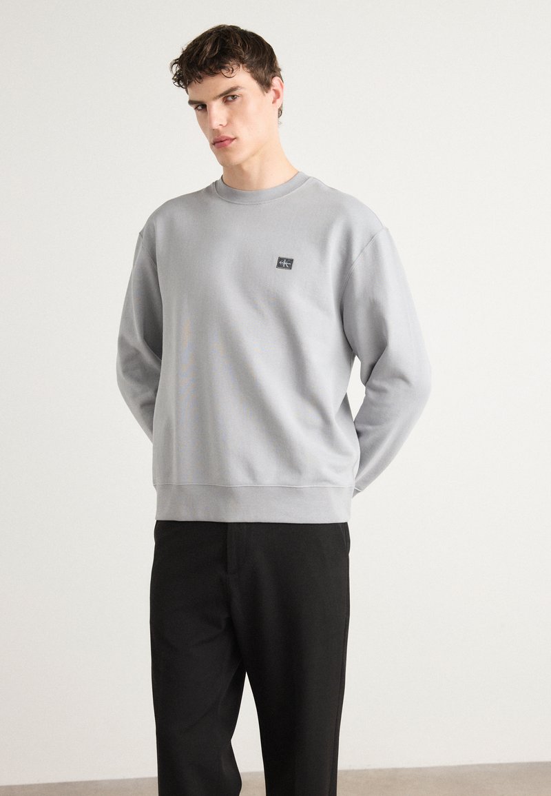 Sudadera de algodón gris con cuello redondo, puños y dobladillo acanalados, que presenta un pequeño parche de logo negro en el pecho. Combinada con pantalones negros.