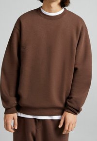 Bruine sweatshirt gemaakt van fleece materiaal met een ruime pasvorm, ribgebreide manchetten en halslijn. Kenmerkt zich door een subtiele logodetail aan de voorkant.