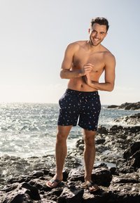 Marine zwemshorts met kleurrijke patronen, met een trekkoord in de taille en een wit logo. Gedragen met zwarte slippers, gepresenteerd tegen een rotsachtige kustlijn.