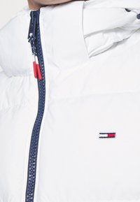 Vit pufferjacka med hög krage, marinblå dragkedja och röda och vita detaljer. Har en liten broderad logotyp på bröstet.