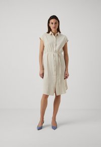 Rochie cămașă de culoare crem, lungime până la genunchi, cu mâneci scurte, deschidere cu nasturi, curea la talie și textură. Purtați cu tocuri ascuțite de culoare bleu deschis.