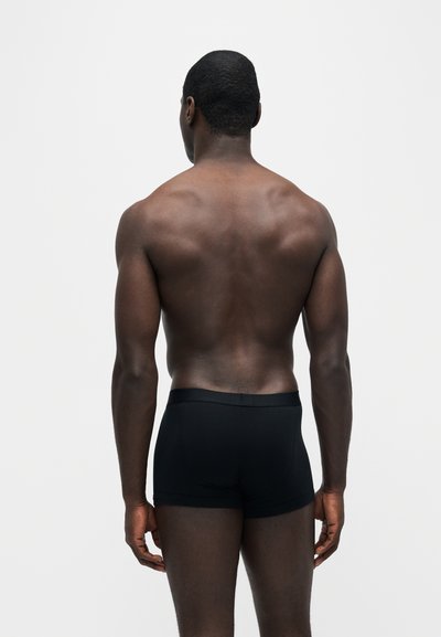 Homme musclé à la peau foncée debout de dos, portant un boxer noir ajusté sur un fond clair uni.