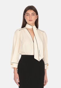 Blusa crema con accenti neri, dotata di colletto con fiocco, lunghe maniche a palloncino e dettagli in oro ai polsini delle maniche, abbinata a una gonna nera.