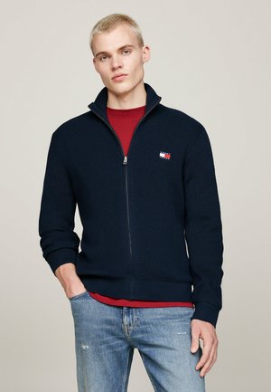 Pull zip-up côtelé bleu marine avec un col montant, orné d'un petit patch logo multicolore sur la poitrine gauche et d'un tissu tricoté texturé.