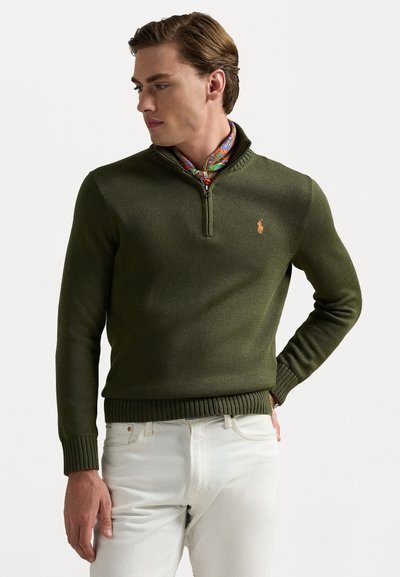 Pulover tricotat de culoare verde măsliniu cu guler tip quarter-zip și tiv ondulat. Prezintă un logo pe piept și este purtat peste o eșarfă colorată cu model.