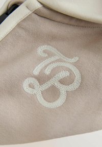 Beżowy materiał o miękkiej fakturze, z wyraźnym białym haftowanym logo w eleganckim, płynnym stylu pisma.