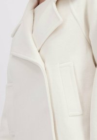 Close-up van een crèmekleurige wolkenjas met brede revers en een rechthoekige patchzak aan de voorkant.