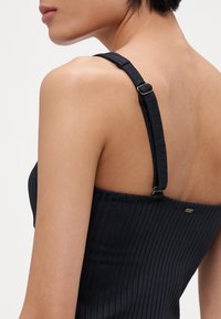 Black ribbed tank top featuring adjustable straps with a metal buckle and a small logo tag on the back. Smooth texture.