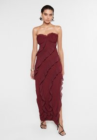 Burgundy strapless mekko, joka on valmistettu tekstuurisesta kankaasta, ja siinä on kerrokselliset rypytykset sekä sivuaukko. Korostettu herkällä rypytetyllä reunuksella.