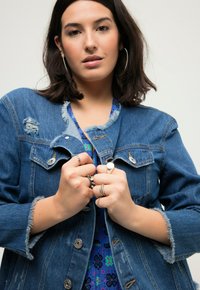 Chaqueta de denim con detalles desgastados, botones plateados y puños deshilachados, llevada sobre una blusa azul estampada. Las manos se entrelazan en la parte frontal de la chaqueta.