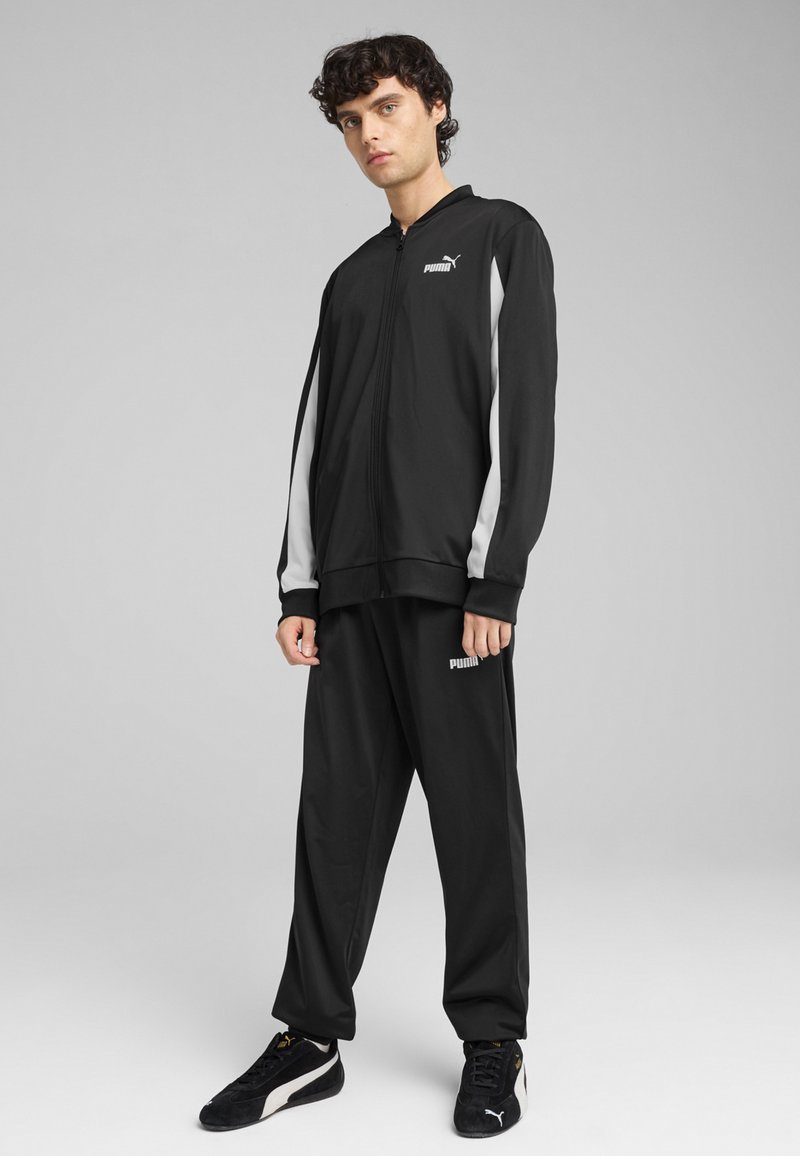 Schwarzer sportlicher Trainingsanzug aus glattem Stoff, mit weißen Akzenten an den Ärmeln und einem kleinen Logo auf der Brust und den Hosen. Strukturiertes Sneaker-Modell.
