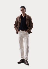 Brun ruskindsjakke over en sort polo, parret med hvide jeans og brune snørresko. Modellen står med hænderne i lommerne mod en lys baggrund.