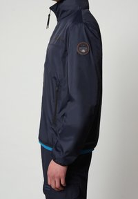 Giacca antipioggia navy con colletto alto, tasca laterale con zip e una patch testurizzata sul braccio sinistro. Materiale liscio e leggero.