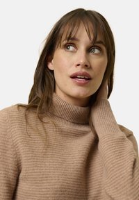 Maglione beige a collo alto con texture morbida. Presenta maniche lunghe e un colletto aderente. Indossato in modo informale con capelli sciolti.