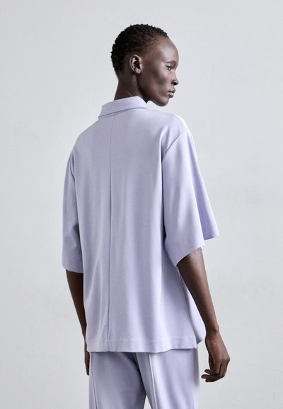 Lavender short-sleeve polo shirt featuring a back seam, a flat collar, and a relaxed fit, made from smooth fabric with a slight sheen.