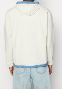 Ljus crème jacka med blå huva, elastiska muddar och blå nederkant. Har en mjuk textur och enkel design.