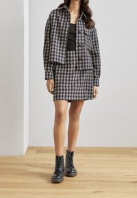 Chaqueta de franela a cuadros en azul marino y gris, combinada con una falda a juego. La modelo lleva botas negras de cordones. Fondo de suelo de madera.