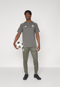 Grå sporty skjorte med korte ærmer og logo, parret med matchende tapered bukser med grønne accenter. Modellen holder en fodbold.