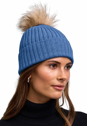 Giovane donna che indossa un berretto a coste blu con un grande pon pon in pelliccia sintetica marrone chiaro e un maglione nero a collo alto.