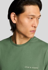 Grøn bomuld T-shirt med rund hals og et lille hvidt "LYLE & SCOTT" logo på venstre side af brystet. Glat tekstur.