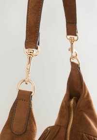 Lanière de sac en daim marron avec boucles métalliques dorées et détail de fermeture éclair sur le côté du sac visible sur un fond clair.