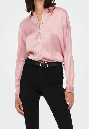 Femme portant un chemisier rose brillant boutonné, rentré dans un pantalon noir taille haute avec une ceinture noire avec une boucle dorée.