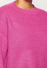 Rosa stickad tröja med ribbad textur, rund halsringning och sänkt axelsöm. Materialet verkar mjukt och varmt.