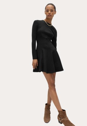 Kobieta w dopasowanej czarnej sukience z długim rękawem i rozkloszowanym dołem, brązowych botkach za kostkę oraz złotym łańcuszkiem na gładkim tle.