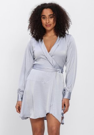 Vrouw met krullend haar, gekleed in een lichtgrijze satijnen wikkeljurk met lange mouwen, een asymmetrische zoom en een taillestrik.