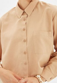 Camicia beige con bottoni, caratterizzata da un colletto classico, taschino frontale e tessuto morbido. I bottoni sono dello stesso colore della camicia, con una vestibilità morbida e maniche con polsini.