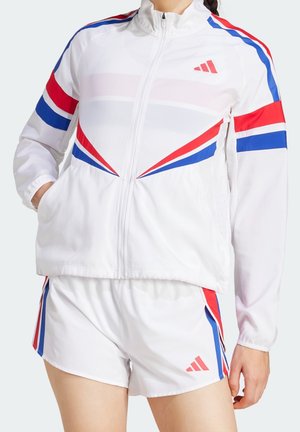 Witte sportjas en korte broek met rode en blauwe diagonale strepen en Adidas-logo's, gedragen door een persoon met handen in de zakken.
