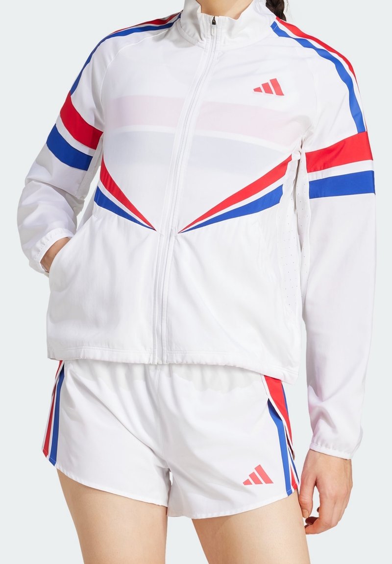 Veste de sport blanche et short avec des rayures diagonales rouges et bleues et des logos Adidas, portés par une personne les mains dans les poches.