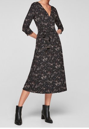 Kobieta ubrana w czarną sukienkę midi w biało-różowy wzór kwiatowy, z rękawami o długości 3/4, dekoltem w kształcie litery V, przewiązaną talią i czarnymi botkami do kostek.