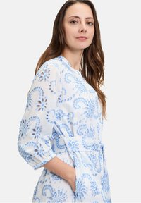 Abito bianco con ricami azzurri chiari che presentano motivi floreali, maniche lunghe, una cintura in vita e una vestibilità comoda.