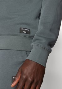 Felpa grigia con polsini e orlo a coste, caratterizzata da un'etichetta nera con testo bianco che descrive il marchio e l'origine, abbinata a pantaloni coordinati.