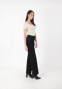 Top corto beige e pantaloni neri a vita alta con cuciture verticali. Il modello indossa stivaletti neri con tacco. Lo sfondo è bianco.