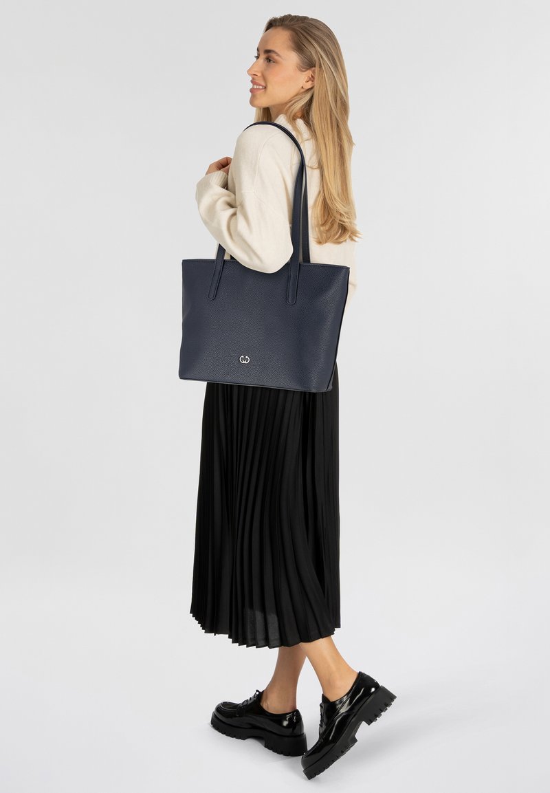 Borsa a tracolla in tessuto blu navy con due manici lunghi, con un logo sul fronte, abbinata a una gonna plissettata nera e scarpe nere lucide.