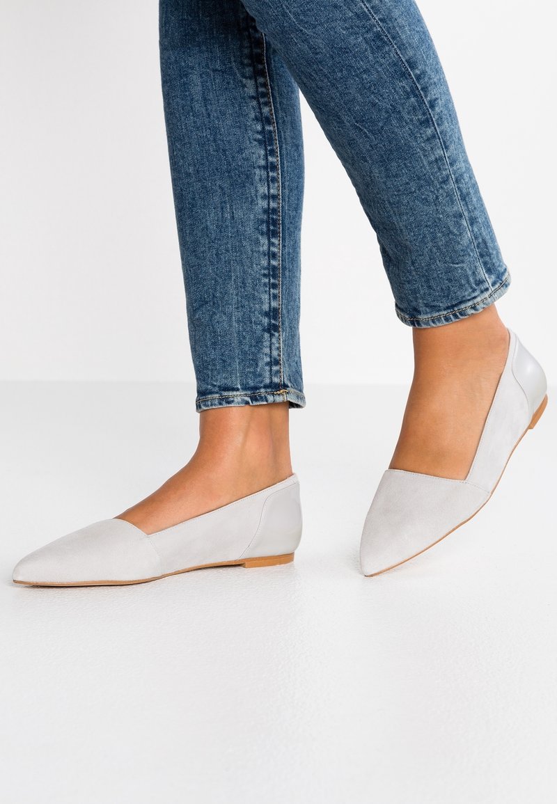 Personne portant des ballerines gris clair à bout pointu avec de petits talons, associées à un jean en denim bleu, debout sur un sol blanc.