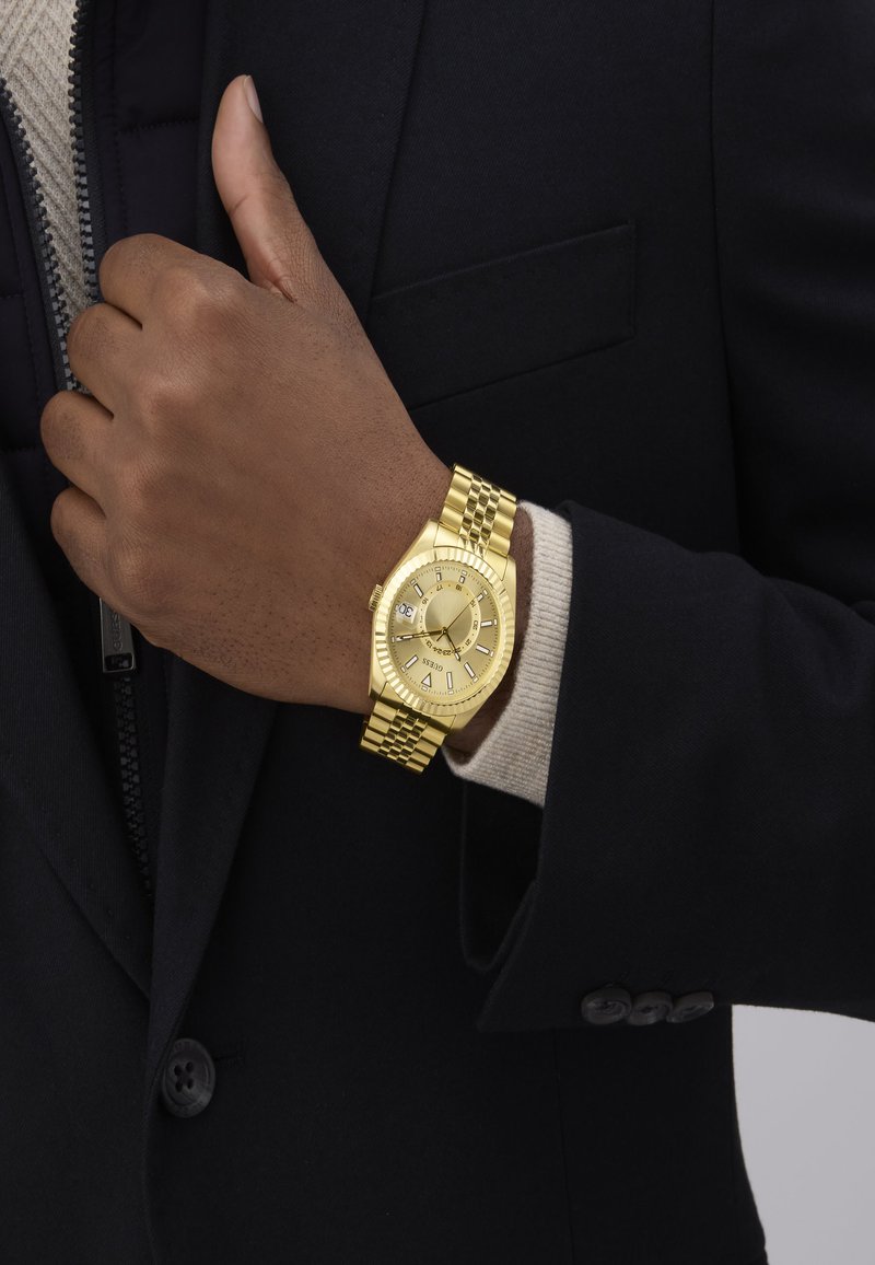 Gul armbåndsur med en rund urskive, tekstureret bezel og en sammenkoblet rem. Hånden viser et mørkt jakkesæt og en lys sweater under.