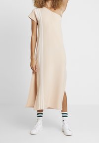 Beige šaty na jedno rameno se skluzem na boku, s bílými pruhy podél levé strany. Vyrobeno z hladkého materiálu, v kombinaci s bílými teniskami a pruhovanými ponožkami.