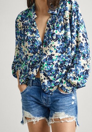 Kvinde iført en blå blomstret bluse med lange ærmer stoppet ned i slidte blå denimshorts med hænderne i lommerne.